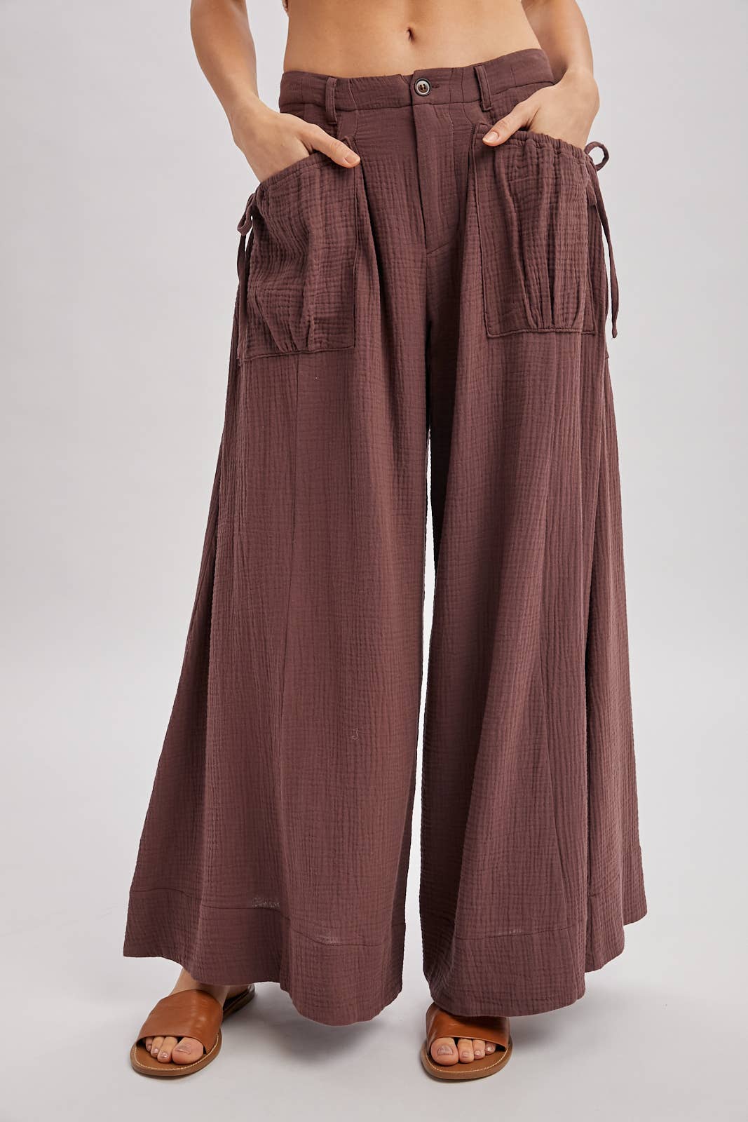 Women's brown gauze wide leg pants, large front pockets, side tie pocket details, belt loops, zip up, button closure, paired with brown sandals.