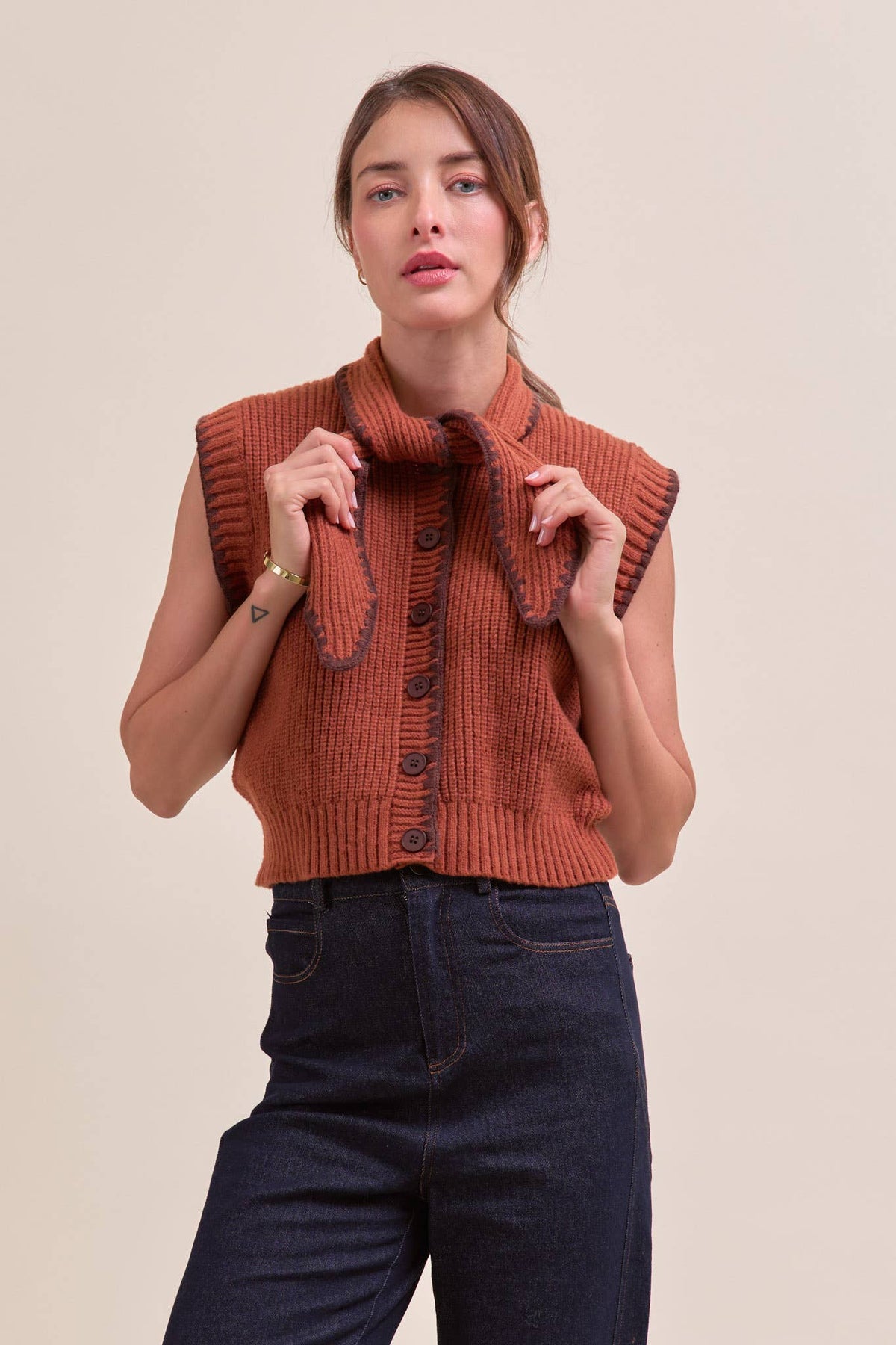 Women's rust-orange ribbed knit sleeveless button-front top with a high folded tie neck scarf collar, dark brown trim detail, paired with dark blue jeans. 