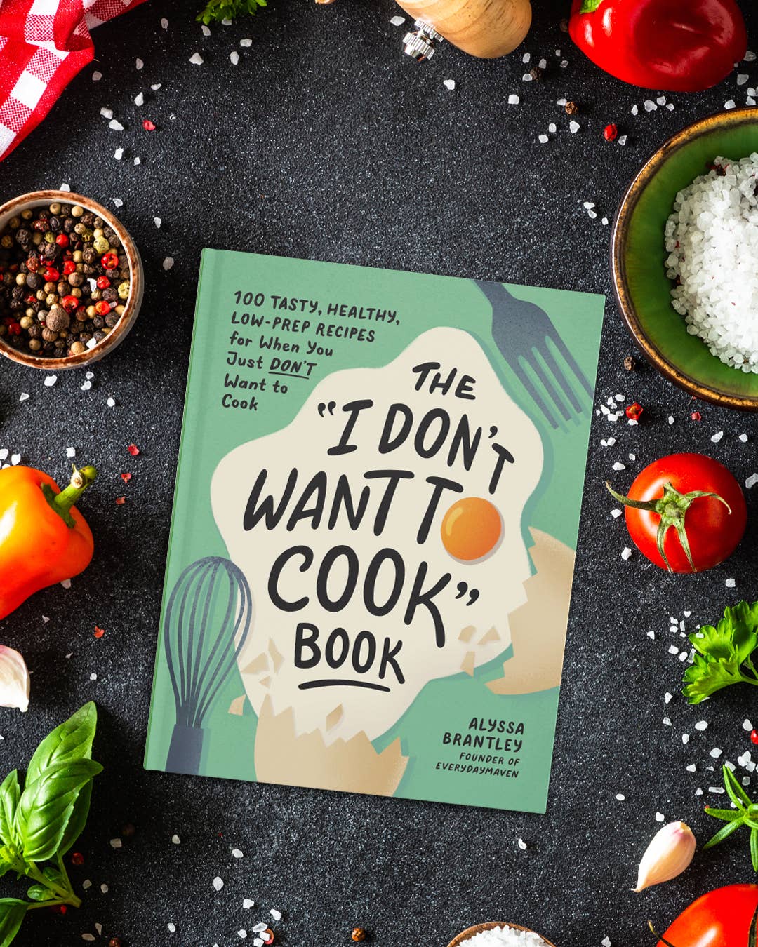Book titled 'I Don't Want to Cook' surrounded by kitchen ingredients on a dark surface, 100 15 minute or less recipes 
