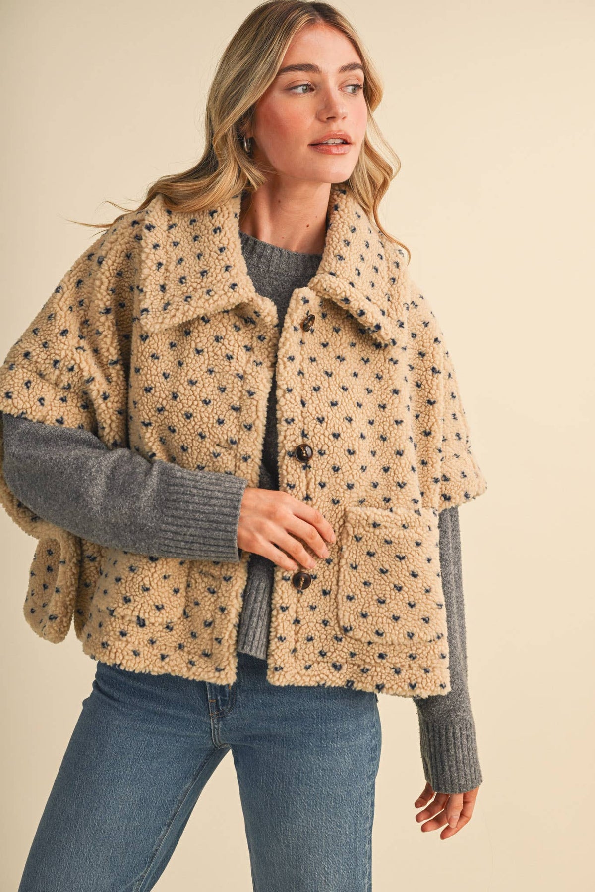 Women's sherpa boyfriend jacket in a warm taupe blue heart printed detail fabric, side button closure, short sleeve, coverts to a poncho, button front, paired with a grey sweater and jeans. 
